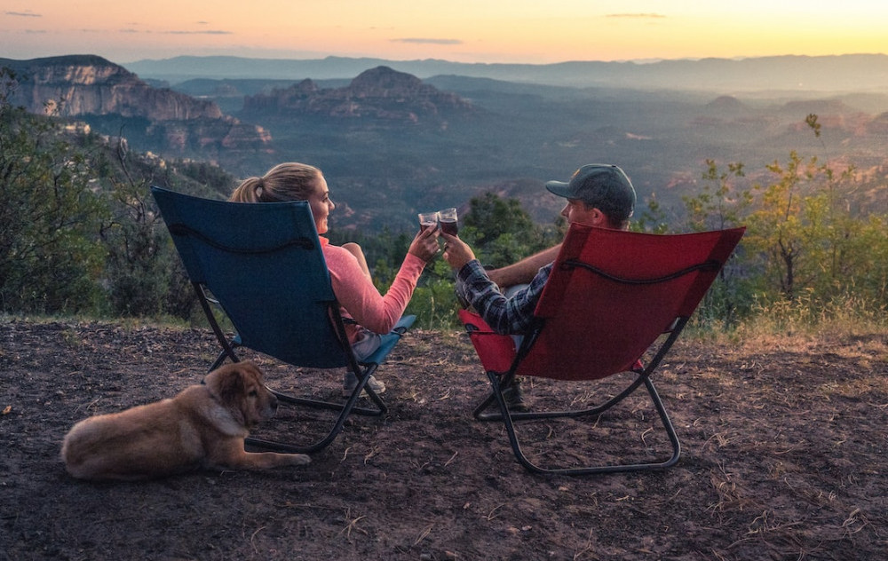 A couple sitting in camping chairs drinking a glass of wine overlooking the valley below while their dog lays behind them resting