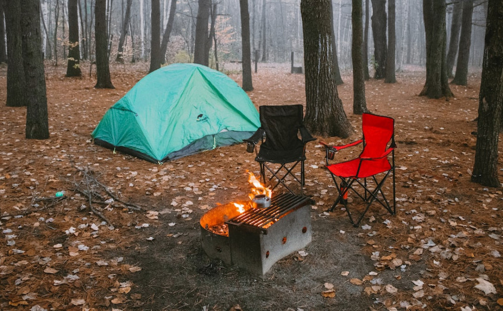 Two empty camping chairs around a firepit with a green tent behind in the middle of woodland