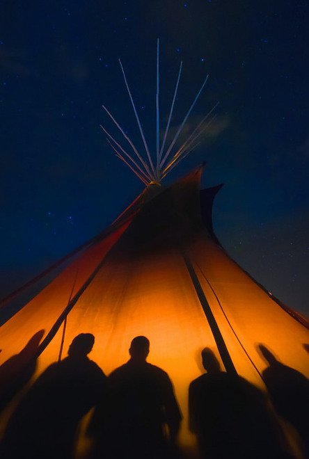 Picture of six people sat in a Teepee at night with smoke coming from the top of the Teepee surrounded by a warm glow