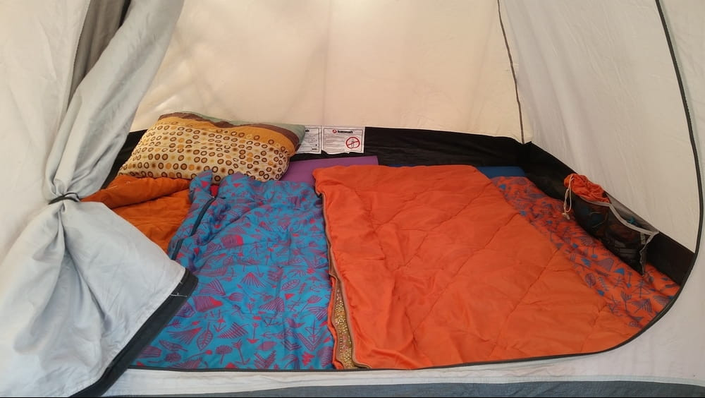 A white open tent showing a pillow and a blue/red sleeping bag and an orange sleeping bag