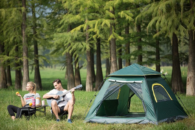 Couple camping near woods sat outside on chairs, the man playing a guitar and the woman sat listening eating fruit