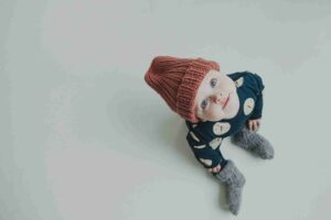 Baby wearing a red hat, green jumpsuit and grey woollen socks looking up at the camera