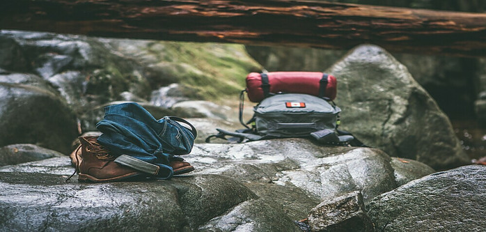 Two backpacks and a pair of hiking boots placed on a rock under a dead fallen tree