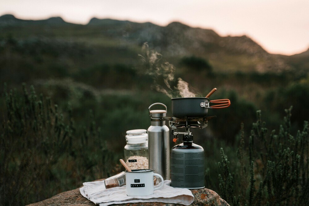 Camping pot placed on a camping stove smoking away with a flask and cup near by placed on a cloth