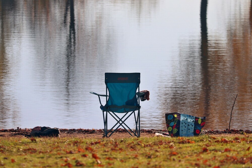 Empty camping chair placed by a lake with a large bag beside it