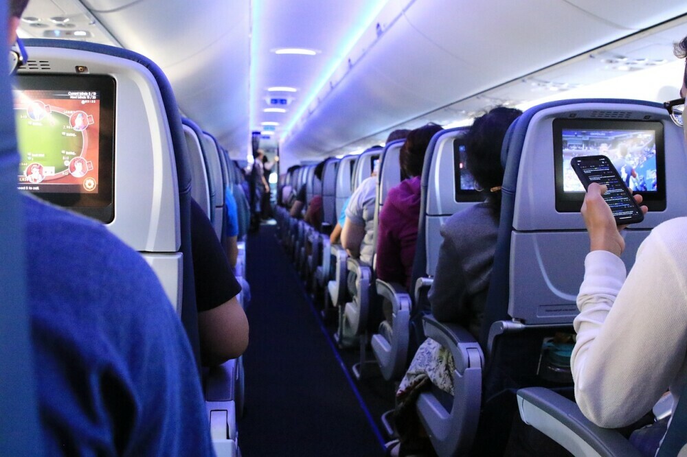 View of passengers on a plane from behind looking forward toward the front with a view of all the monitors on the back of their seats