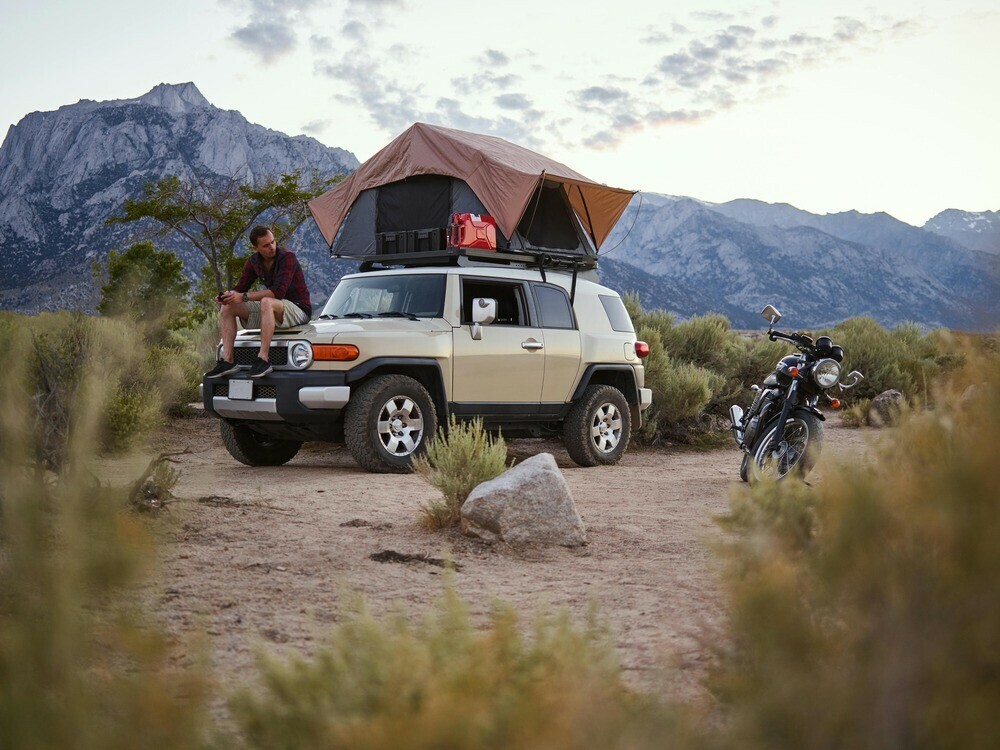 Guy sat on the hood of his vehicle with his roof-top tent setup and a motorcycle close by parked in the desert with mountains in the background